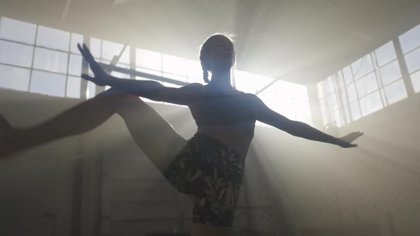 A female performs dance moves in a large dark room with sunlight beaming in the window