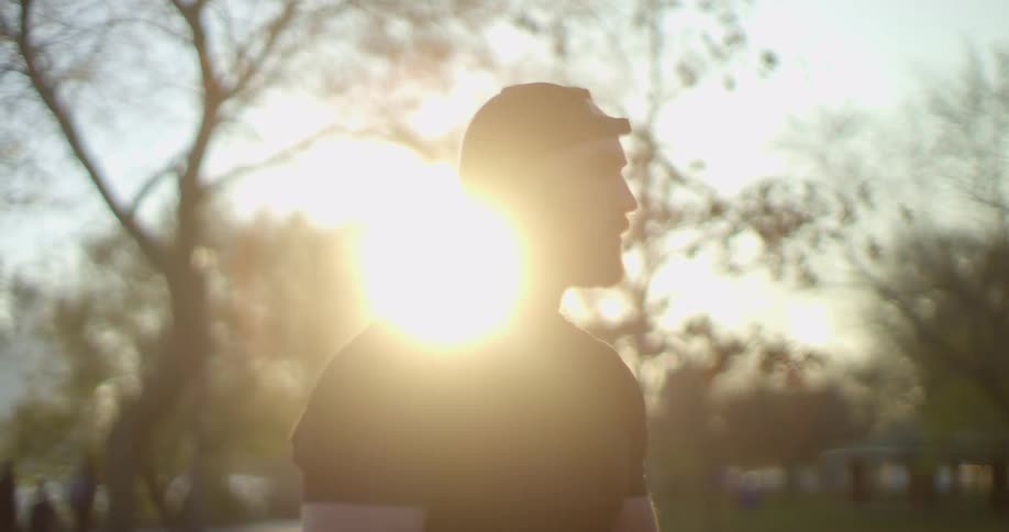 Young bearded man wearing baseball hat standing on a park at sunset