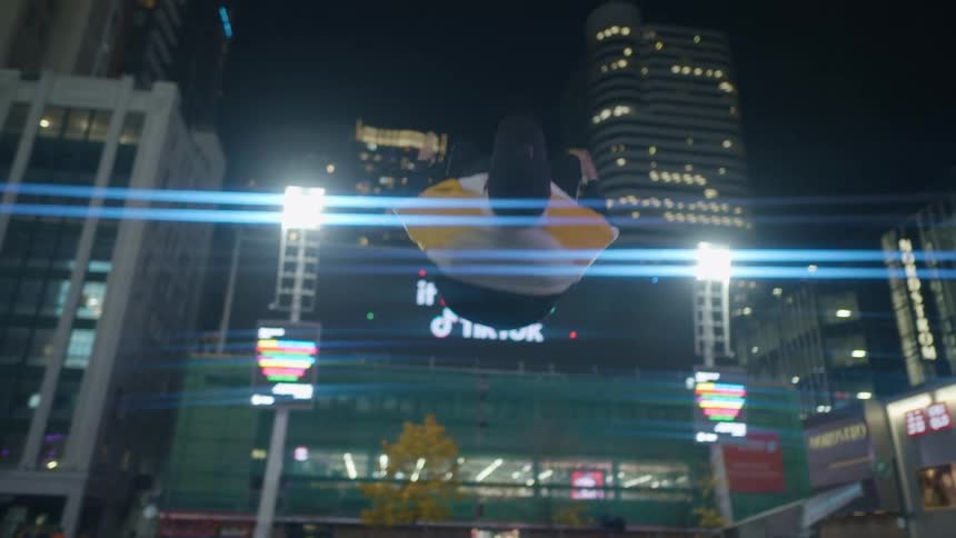 A man standing in a plaza surrounded by tall buildings performs a standing back flip