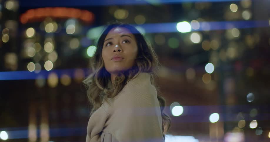 A woman stands downtown at night and looks up at the brightly lit buildings