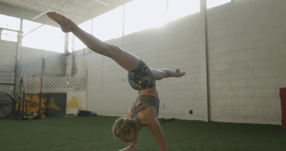 A woman in a room stretching with a man jumping and flipping over her