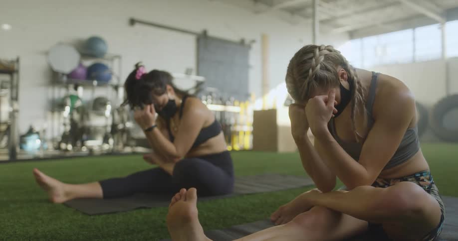 Two women sit on yoga mats in a gym area doing yoga and meditation
