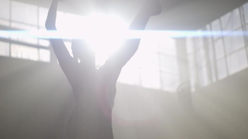 A female stands in a large room lifting her leg in a dance move with bright sunlight beaming inside