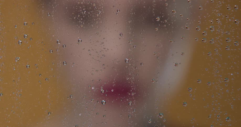 Young woman with red lipstick smoking a cigarette behind a wet glass pane