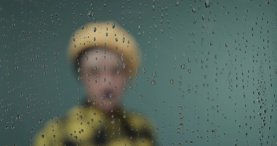 A person in a yellow hat and coat stands behind a glass with water droplets