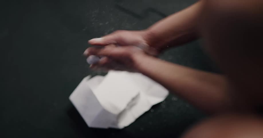 A person kneeling in a gym covers their hands in athletic chalk before clapping their hands together