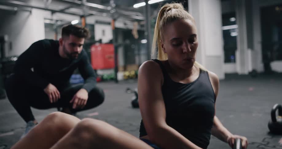 A woman works out with weights in a gym as a male trainer is next to her