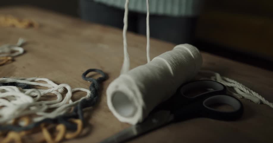 A woman unrolls white string from a spool on a craft table next to scissors and other string in different colors