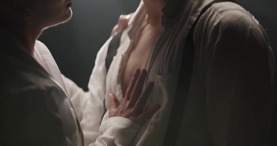 A woman runs her hand down the chest of a man as they dance together under a light in an otherwise dark room