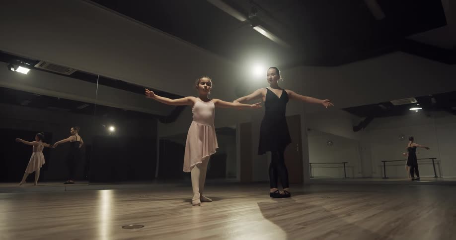 A girl practices ballet poses under the instruction of a woman in a ballet studio