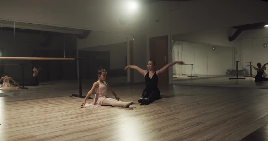 A ballet instructor leads a girl in performing ballet poses on the floor of a ballet studio