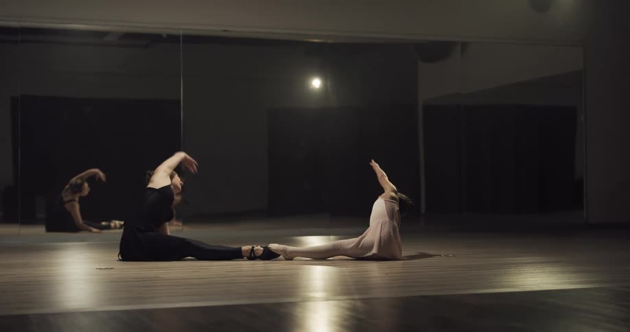 A young girl sits on the ground with her feet touching her ballet teachers feet as they mimic stretching together