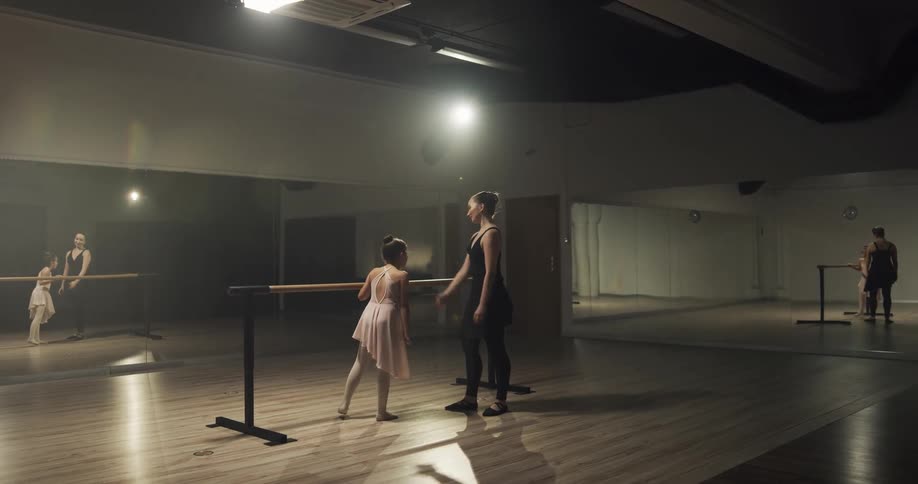 A dance teacher helps a young girl stretch and hold a dance pose