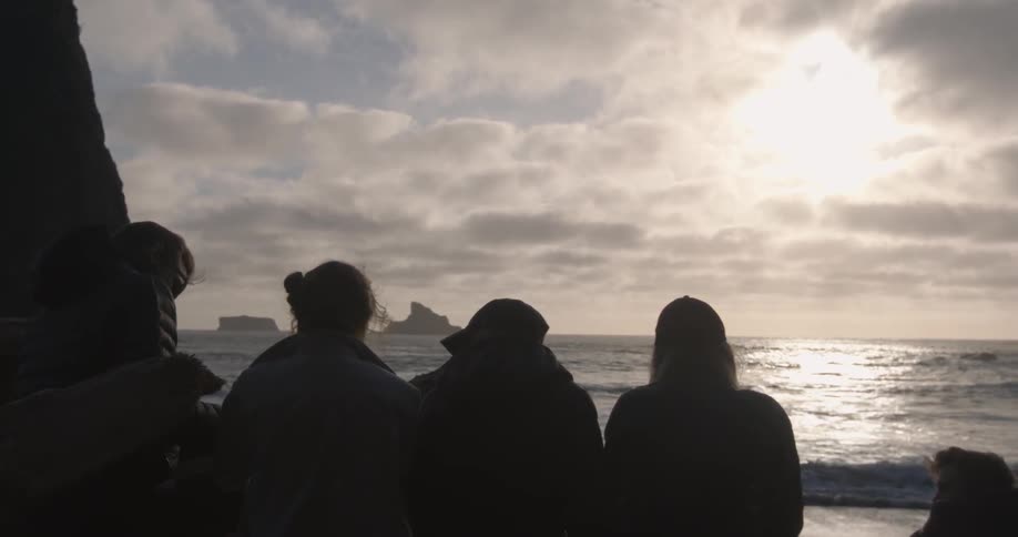 Several people looking towards the waters at a beach with the sun shining through the clouds