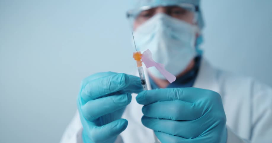 A doctor in a face mask and white lab coat holding a syringe