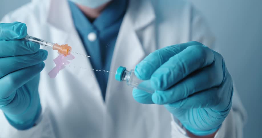A person in a white lab coat pulls a needle out of a syringe and liquid sprays out