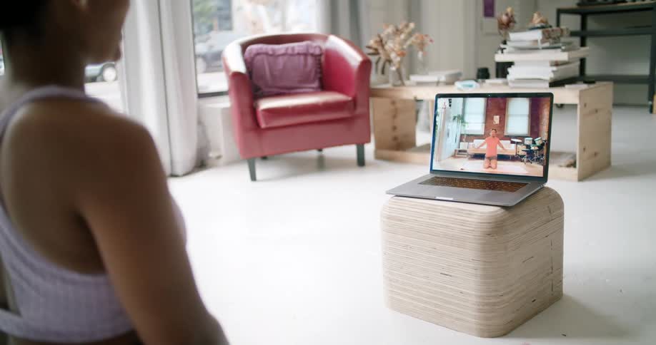 A woman sits in a living room exercising with an instructor on a laptop