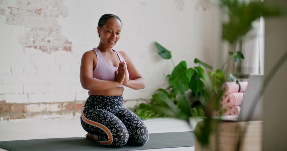 A woman kneels on a yoga mat and bows in front of an open laptop