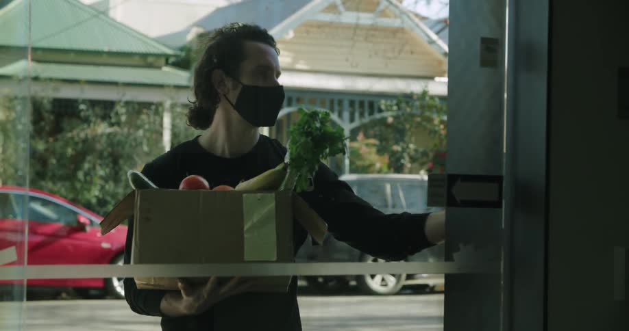 A man delivering a box of food to a house looks at his cell phone and then rings the doorbell