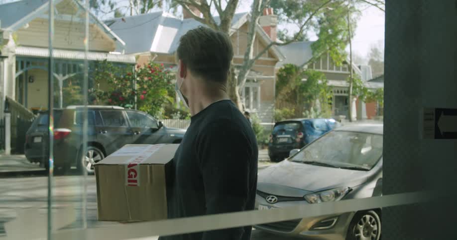 A man with a mask and a smart phone walking up to a building and handing a box to a person