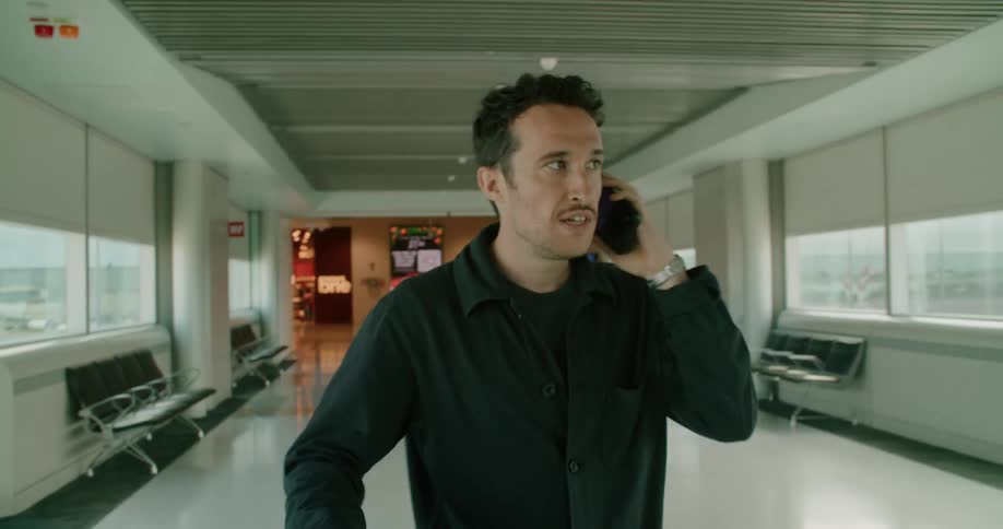 A man walking in an airport and talking on a smart phone with chairs and windows in the background