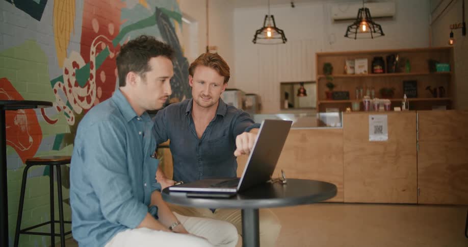 Two men sit in a shop and look at a laptop