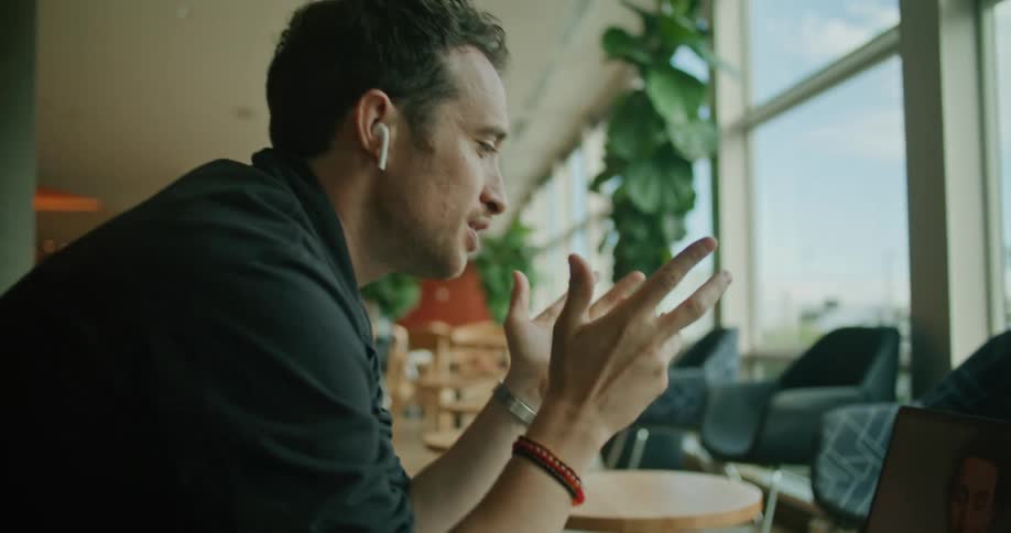 A man sits at a small table in an empty cafe and talks to another man on his laptop via video call