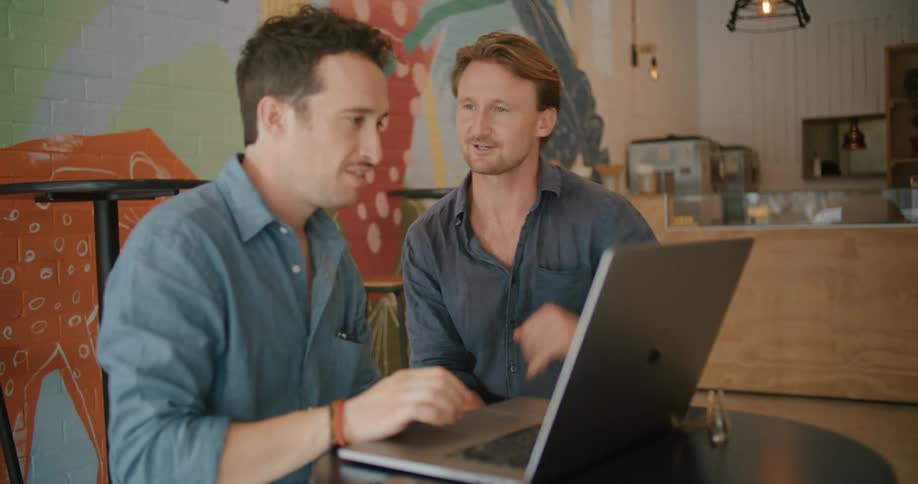 Two men work on a laptop together while sitting at a small table inside a cafe