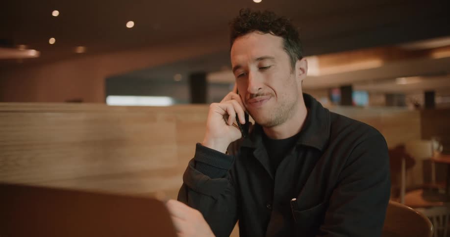 A man talks on his smartphone while using a laptop at a table in a restaurant