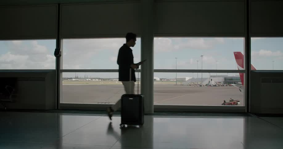 A man with a rolling suitcase walks through an airport where he passes a large window