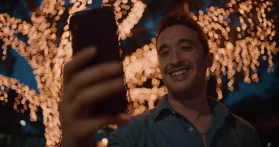 A man standing outside in front of a tree lit up with lights video chats on his cell phone
