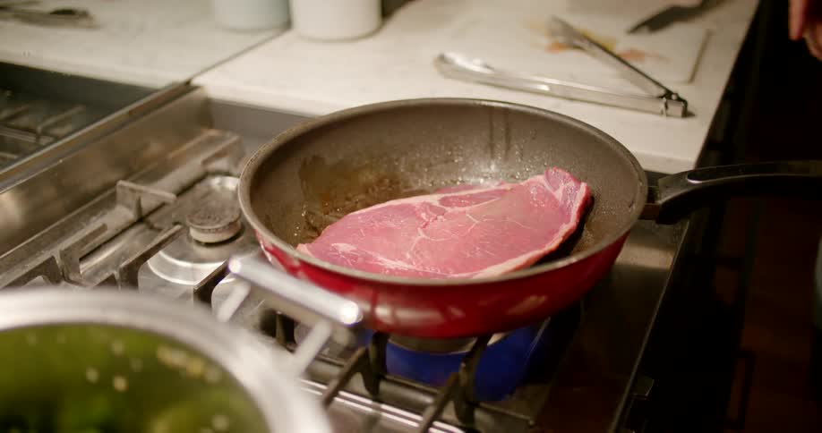 A person putting meat into a frying pan on a stove with fire