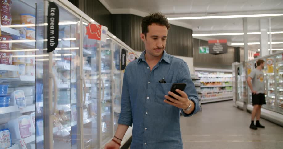 A man getting something out of a refrigerator at a store and staring at his phone