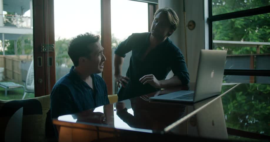 A man leans against a table with a laptop on it talking to a seated man