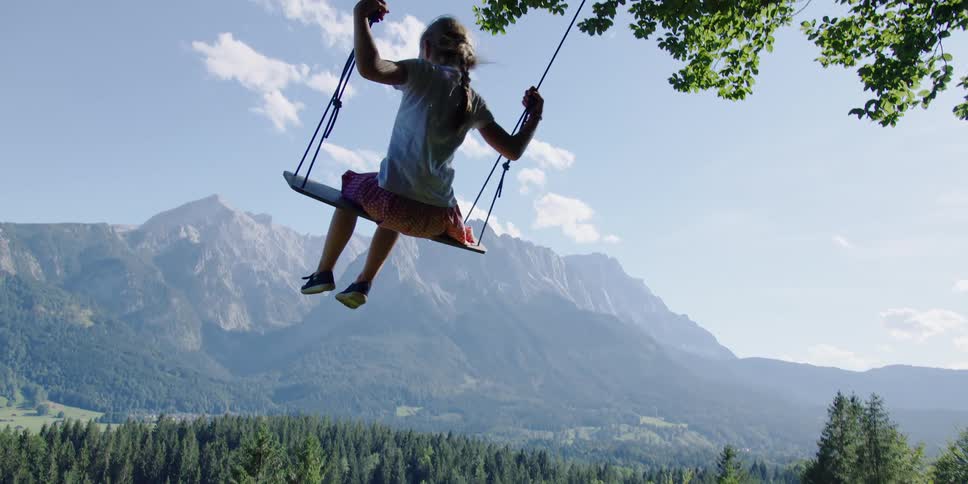 A girl swings on a swingset on a hill overlooking a forested countryside below a mountain ridge on a sunny day