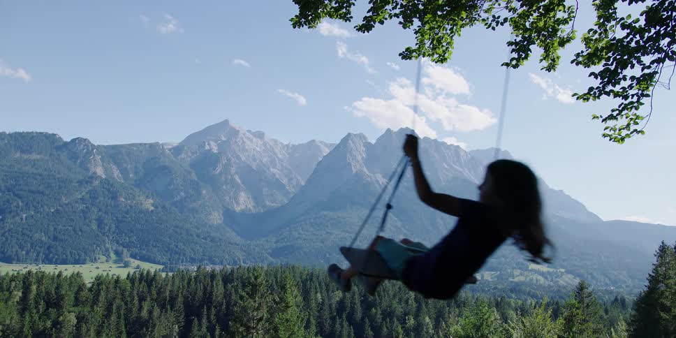 Young girl playing in a swing hanging from a tree in front of the mountains