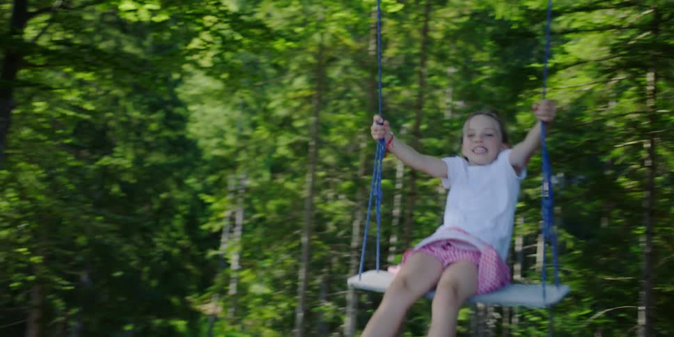 A girl laughs and smiles as she rides a swing hanging from a tree at the edge of a forest overlooking a mountain range
