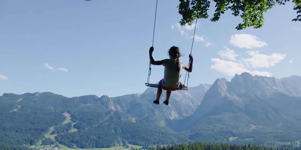 A young girl holds on tight while riding a swing hanging from a tree near a mountain range