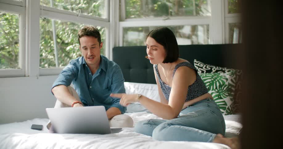 A man and woman are sitting on a bed and looking at a computer