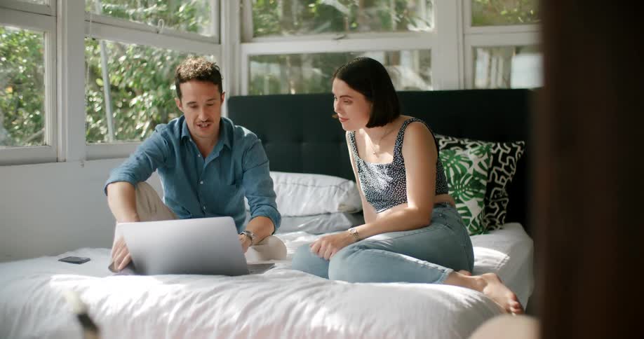 A man and woman are sitting on a bed next to a laptop