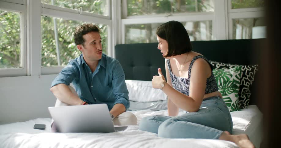 A woman and man are sitting on their bed with a laptop