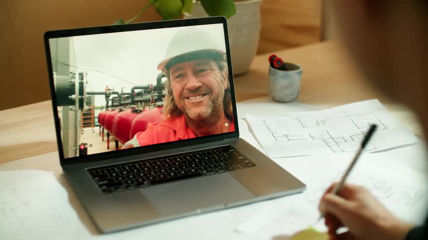 A man in an office makes some notes as he talks to a coworker in a hardhat on video call on a laptop