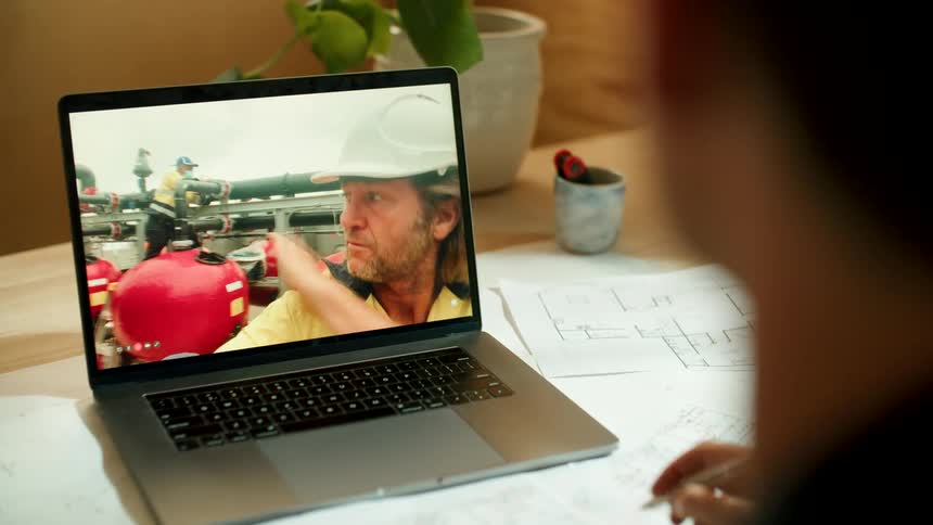 A architect sits at his desk with blueprints while talking via video call to a colleague at a construction site