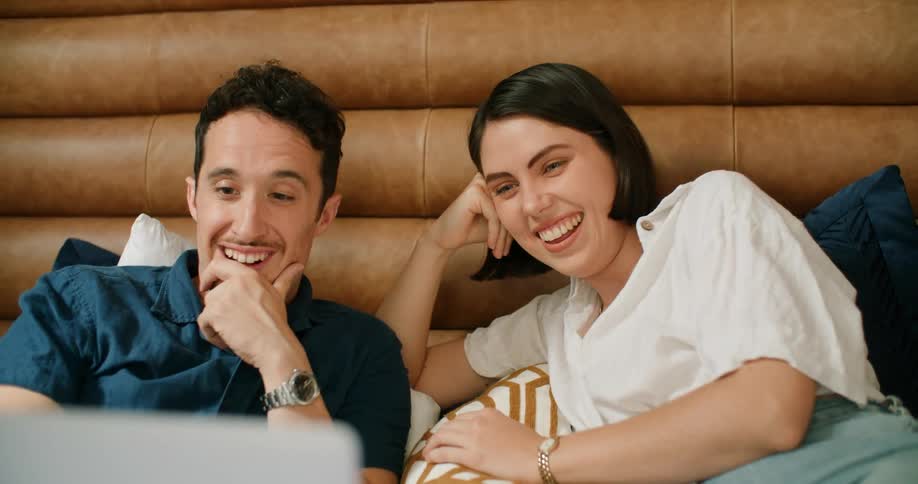A man and woman laugh while laying in bed and watching a video on a laptop