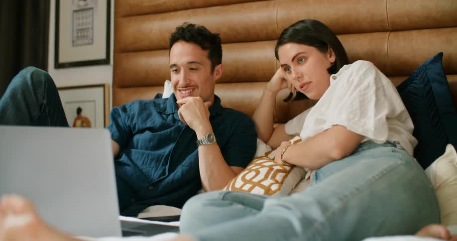 A husband and wife smiling while watching a movie on a laptop in bed