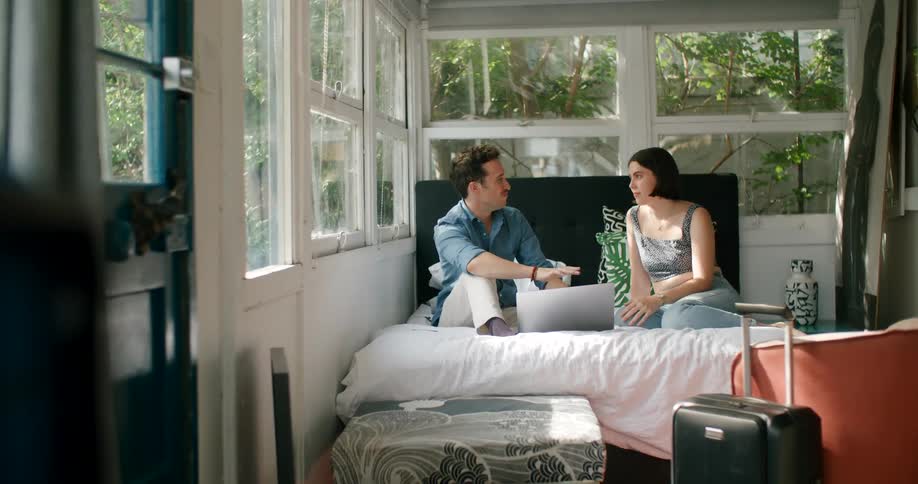 A man and woman sit and talk in bed at a cottage surrounded by trees