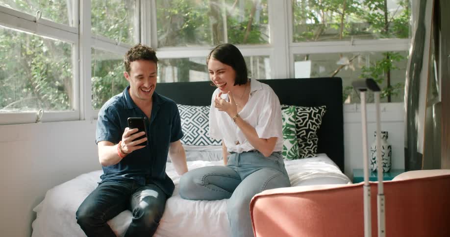 A man and woman sitting side by side on a bed laugh while gazing at the screen of a smartphone