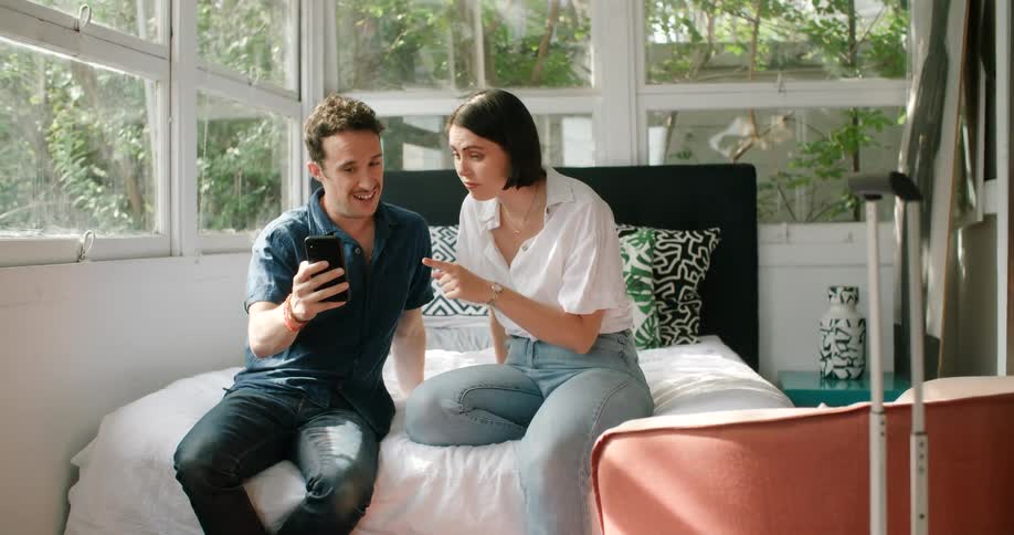 A man and woman sit side by side on a bed smiling and talking on a phone