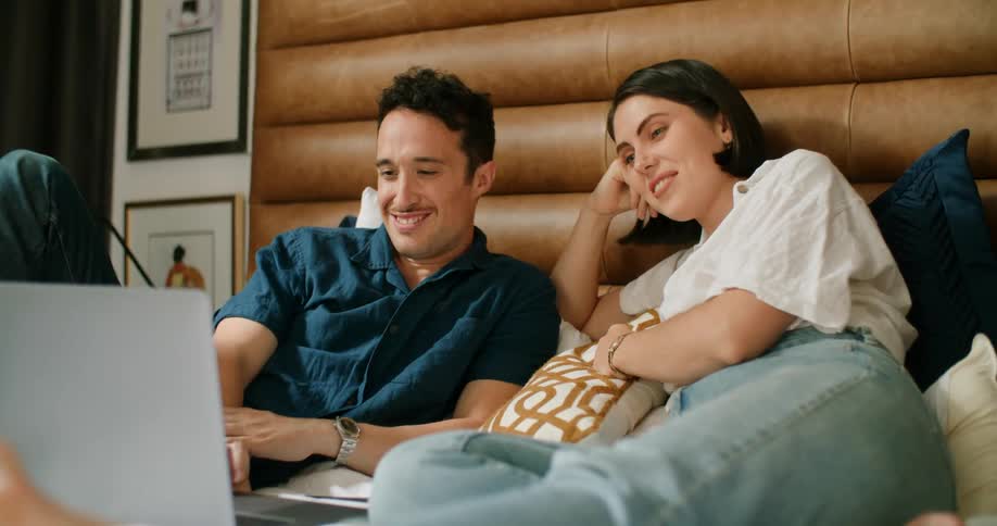 A man and woman laugh as they lie in bed and watch a video on a laptop