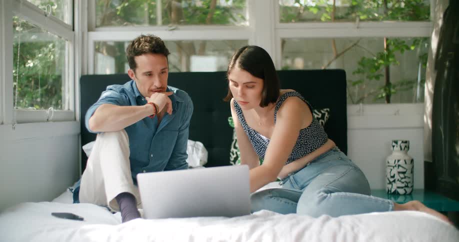 A man and woman sit on a bed and look at a laptop